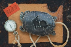 a closeup vignette of a tortoise with a rabbit stencil on its back atop a rough edged plank with stopwatch in frame. A card states “Ten hops, Uncle Wiggily! My, but that’s fine, Some rabbits I know would only get nine”