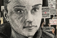 A portrait of a young activist, the woman holds sharp eye contact with the viewer. The background shows signs of protest saying "Girl Power", "Women's Rights = Human Rights", "equality", etc.