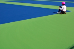 image of a woman sitting on a skateboard, with birght pink helmet, blue and green surface of the ground.